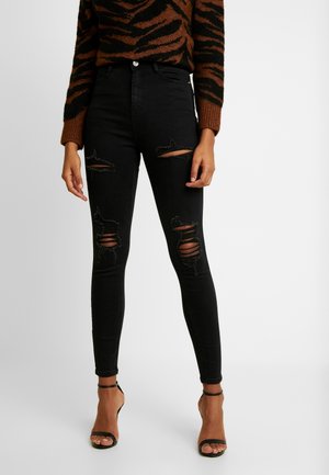 Kobieta ubrana w czarne obcisłe podarte jeansy z wysokim stanem, brązowo-czarny sweter w tygrysie paski oraz czarne sandały na obcasie z odkrytymi palcami i paskiem wokół kostki, stoi.