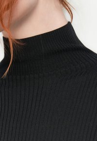 Must mustriba turtleneck-sviiter mustas, millel on kõrge krae ja tekstureeritud disain, rõhutades keha lähedast siluetti ja pehmet kudumit.