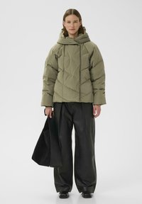 Giacca a vento verde oliva con cappuccio, caratterizzata da un motivo trapuntato e cordini, abbinata a pantaloni larghi in pelle nera.