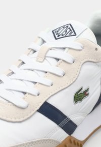 Witte sneaker met beige en marineblauwe suède accenten, voorzien van een groen krokodillenlogo, witte veters en een gestructureerde rubberen zool.