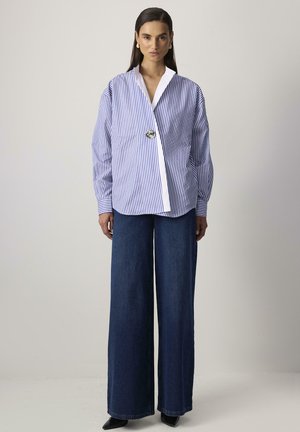 Woman wearing oversized blue and white striped blouse with a large button and wide-leg dark blue jeans, standing against a plain background.