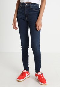 Pantalones vaqueros ajustados de tiro alto en azul oscuro, hechos de denim, con un diseño clásico de cinco bolsillos y combinados con zapatillas rojas brillantes.