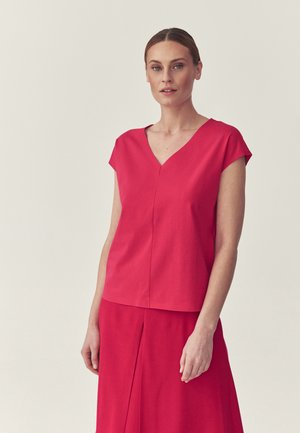 Woman with light skin and tied-back hair wearing a red short-sleeve V-neck top and matching red skirt against a light background.