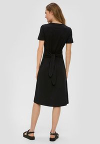 Robe noire à manches courtes avec un détail noué à l'arrière ; longueur genoux, fabriquée dans un tissu lisse avec une coupe décontractée. Des sandales noires complètent le look.