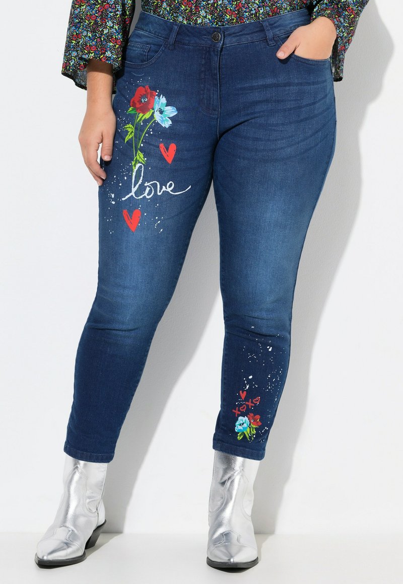 Jeans de mezclilla azul con bordados florales en rojo, turquesa y corazones. Los acentos incluyen botas metálicas plateadas hasta el tobillo y detalles salpicados.