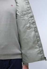 Maglione grigio chiaro con orlo a coste, indossato sotto una giacca imbottita verde chiaro con chiusura a zip e tasca laterale.