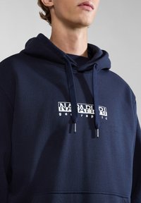 Persona joven vistiendo una sudadera azul marino con el logo blanco "Napapijri geographic" y cordones, mostrada de la barbilla al pecho.