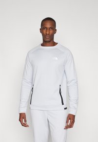 Sweatshirt cinza claro com punhos canelados, apresentando bolsos laterais com fecho éclair pretos e um pequeno logo refletor no peito. Acabamento de tecido suave.
