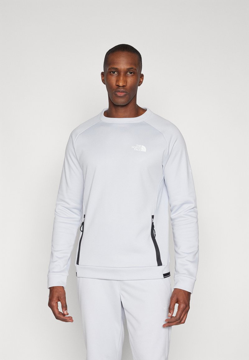 Sweatshirt cinza claro com punhos canelados, apresentando bolsos laterais com fecho éclair pretos e um pequeno logo refletor no peito. Acabamento de tecido suave.
