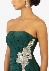 Abito senza spalle di color verde smeraldo con corpino plissettato. Presenta un'applicazione floreale argentata su un lato e orecchini coordinati di grande impatto.