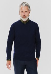 Granatowy sweter w serek z miękkiego materiału, z prążkowanymi mankietami i dołem, noszony na zielonej koszuli z guzikami i kołnierzykiem.
