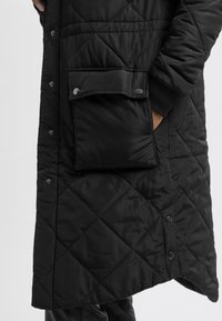 Abrigo negro acolchado con cierre de botones a presión, bolsillo lateral y puños acanalados. El material es suave con un ligero brillo.