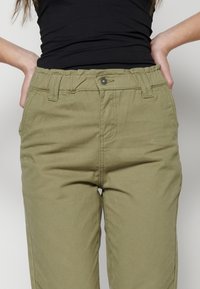 Pantalones verde olivo con un corte relajado, cinturilla elástica y costuras visibles. Presentan bolsillos laterales y un cierre de botón en la parte delantera.