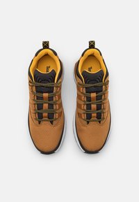 Zapatillas de tela marrones con acentos en negro y amarillo, que presentan una parte superior texturizada, cordones con patrón y una suela de goma blanca.