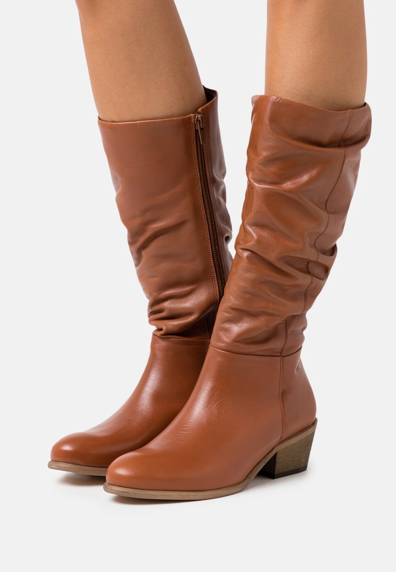 Bottes en cuir marron montant jusqu'au milieu du mollet avec fermeture éclair latérale, tige légèrement froissée, bout arrondi et talon bloc bas, portées sur des jambes nues.