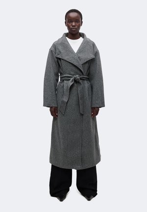 Personne portant un long manteau gris ceinturé par-dessus un haut blanc et un pantalon large noir, debout devant un fond clair uni.