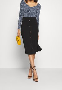 Gonna nera a matita con dettagli dei bottoni frontali, abbinata a una blusa floreale blu a maniche lunghe. Gli accessori includono una piccola borsa gialla.