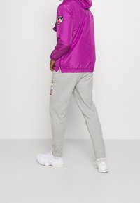Fioletowa nylonowa kurtka z kapturem, z naszywką z logo na rękawie; w zestawie z jasnoszarymi spodniami dresowymi i białymi tenisówkami.