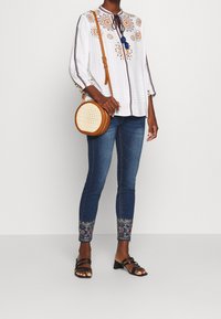 Bílá blůza s barevným květinovým vyšíváním a detailem se zavazováním na krku, kombinovaná s přiléhavými modrými džíny s vyšívanými lemy a černými sandály.