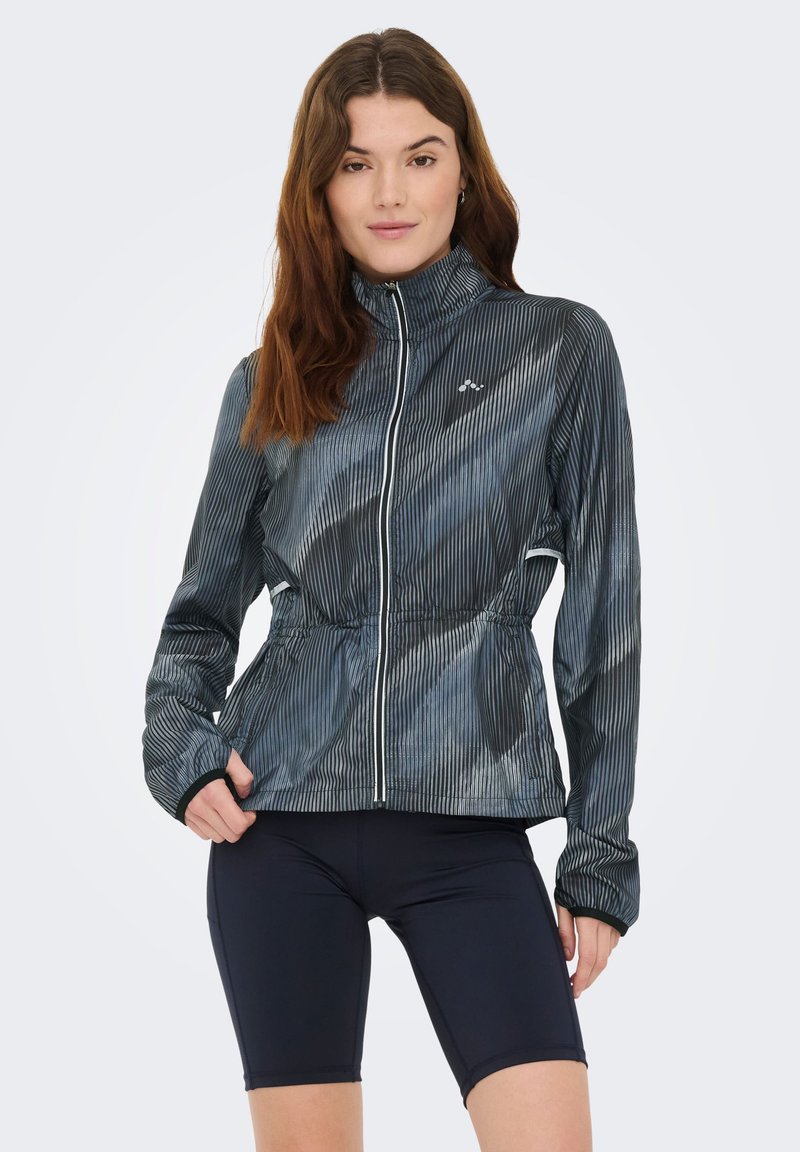 Giacca sportiva blu scuro con strisce verticali, vita elastica, collo alto, cerniera frontale e polsini con foro per il pollice. Abbinata a pantaloncini neri.