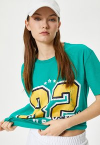 Turkis bomulds t-shirt med et slidt gult "22" design, korte ærmer og en afslappet pasform, brugt med hvide shorts.