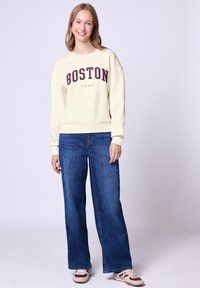 Kołnierzowa bluza w kolorze kości słoniowej z napisem "BOSTON 1980" w bordowym nadruku, połączona z szerokimi niebieskimi dżinsami i wielokolorowymi sneakersami. Gładki materiał, swobodny krój.