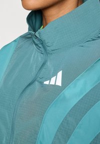 Teal windbreaker s visokim ovratnikom, s zatvaračem i bijelim logom. Tkanina ima teksturirani mrežasti uzorak s glatkim naglascima.