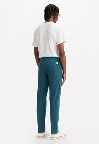 Pantaloni din bumbac turcoaz cu un design drept, cu două buzunare la spate și un mic etichetă de marcă pe talie, purtați cu un tricou alb.