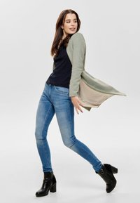 Cardigan lungo verde chiaro sopra un top lavorato a maglia scuro, abbinato a jeans aderenti blu e stivaletti neri con tacco. Tessuto morbido, stile informale.