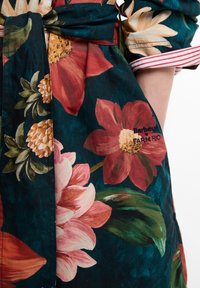 Κοντινή λήψη υφασμάτινης ζώνης με λουλουδάτο σχέδιο σε σκούρο χρώμα, δεμένη στη μέση, με κεντημένο το κείμενο «Barbour Farm Rio» πάνω σε μεγάλο κόκκινο λουλούδι.