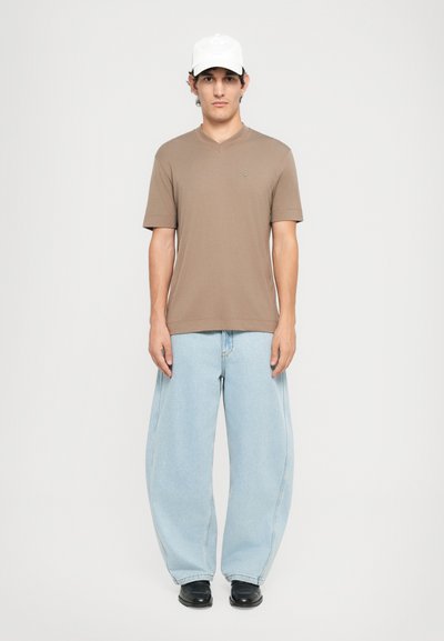 Homme portant une casquette blanche, une chemise à col en V marron à manches courtes, un jean large bleu clair et des chaussures noires, se tenant devant un fond uni.