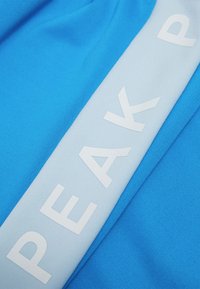 Tessuto atletico blu con una banda orizzontale blu più chiaro che presenta la parola "PEAK" in lettere bianche in grassetto. Texture liscia e colori brillanti.