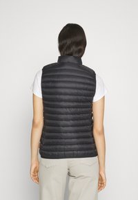 Chaleco acolchado negro con cuello, que presenta una textura acolchada y un diseño ajustado. Lleva encima de una camisa blanca, combinado con pantalones de color claro.