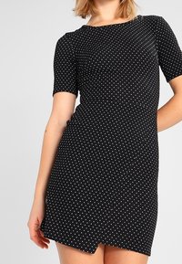 Kortärmad svart klänning med vita prickar, figursydd design, rund halsringning och asymmetrisk nederkant, tillverkad av elastisk tyg.