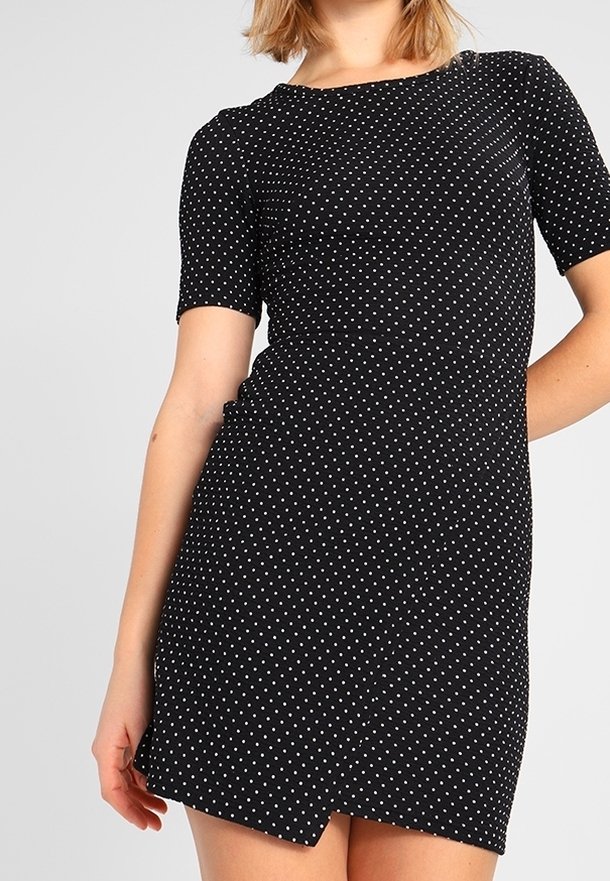Kortärmad svart klänning med vita prickar, figursydd design, rund halsringning och asymmetrisk nederkant, tillverkad av elastisk tyg.