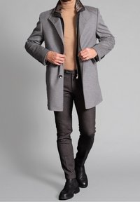 Abrigo de lana gris con cuello de piel, llevado sobre un suéter beige y pantalones gris oscuro. Botines negros completan el conjunto.