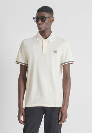 Homme portant un polo écru avec un écusson logo, un pantalon noir et de grandes lunettes de soleil noires, debout devant un fond gris uni.