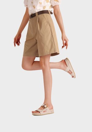 Donna che indossa pantaloncini beige con cintura, sandali bianchi con fibbia tartarugata e una camicia bianca con ricamo floreale giallo, in piedi su una gamba sola.