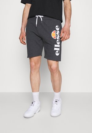 Mężczyzna ubrany w ciemnoszare szorty z troczkiem z dużym białym i pomarańczowym logo „ellesse”, białe skarpety i białe buty sportowe New Balance.
