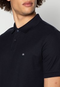 Mörkblå bomullspolo med krage, tvåknappsslutning och broderad 'ck'-logotyp på vänster bröst. Slät textur.