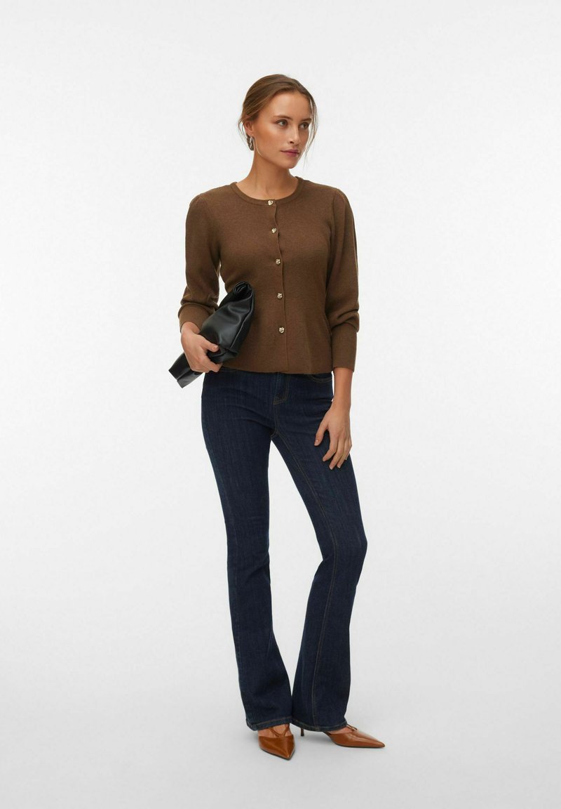 Femme debout portant un cardigan marron boutonné, un jean bleu foncé, des talons marron pointus, tenant une pochette noire.