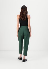 Green, tailored trousers made from a smooth fabric, featuring a high waist, tapered legs, and a rear pocket detail. Black shoes visible.