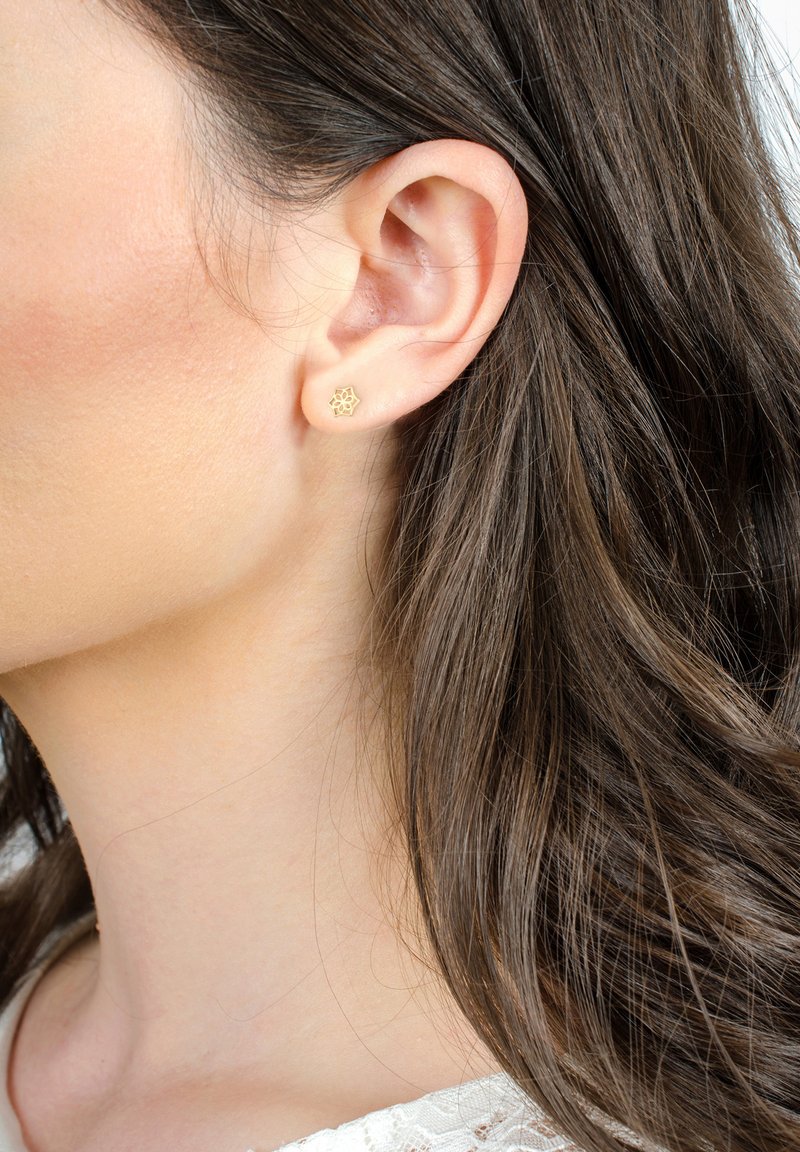 Orecchino a bottone in oro con un intricato disegno floreale, posizionato su uno sfondo di lunghi capelli castano scuro. Tono di pelle naturale visibile.