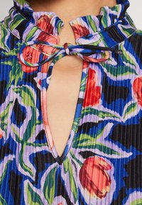 Vestido de estampado floral con un cuello con volantes, cierre atado y un diseño de ojal. La tela presenta pliegues verticales en colores vibrantes.