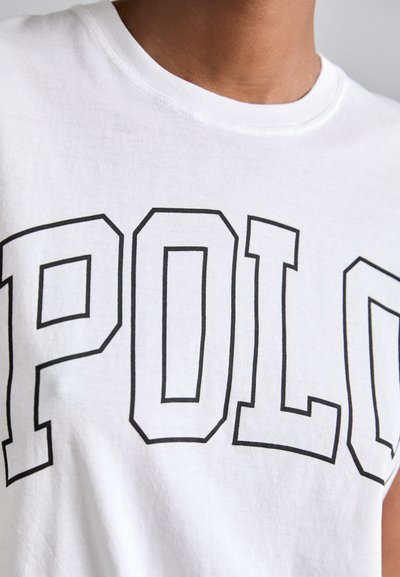 Balta medvilninė marškinėliai su drąsiu juodu kontūru „POLO“ logotipu priekyje. Apvalus kaklas su lygiu paviršiumi ir trumpomis rankovėmis.