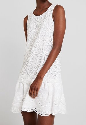Sleeveless white dress with intricate lace patterns on upper and floral embroidery on flared hem, worn by a standing person.