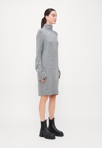 Robe en tricot grise avec un col haut et des manches longues, dotée de poignets côtelés. Associée à des bottes noires à bout chunky.