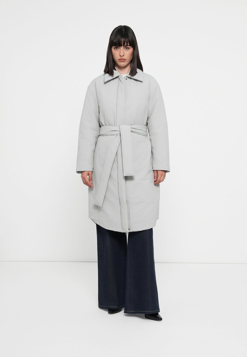 Manteau rembourré gris clair avec un col, taille ceinturée et poches latérales, associé à un pantalon en denim foncé à jambe large.
