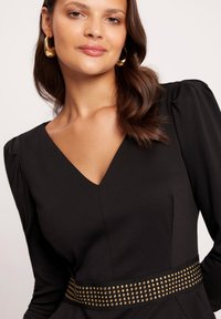 Czarna sukienka z długimi rękawami i głębokim dekoltem w kształcie V, z paskiem ozdobionym złotymi ćwiekami oraz gładkim, teksturowanym materiałem.