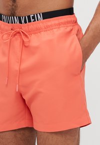Calvin Klein korallfargede badeshorts med elastisk midje med logo, snøring og sidelommer; laget av glatt, lett materiale.